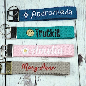 May include: Four fabric keychains with metal hardware. The keychains are teal, pink, gray, and blue. Each keychain has a name embroidered on it: Andromeda, Truckie, Amelia, and Mary Anne. The Truckie keychain has a smiley face.