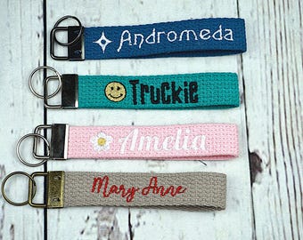 May include: Four fabric keychains with metal hardware. The keychains are teal, pink, gray, and blue. Each keychain has a name embroidered on it: Andromeda, Truckie, Amelia, and Mary Anne. The Truckie keychain has a smiley face.