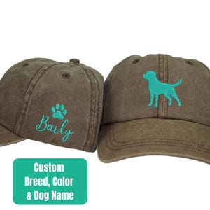 May include: Two brown baseball caps with adjustable straps. One cap has a paw print and the name "Bailey" embroidered in turquoise. The other cap has a turquoise silhouette of a dog embroidered on it. The text "Custom Breed, Color & Dog Name" is printed on a turquoise rectangle.