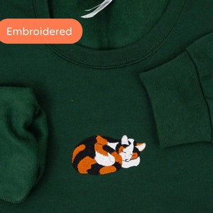 May include: A dark green sweatshirt with an embroidered image of a calico cat sleeping. The sweatshirt has the word "Embroidered" written on it.