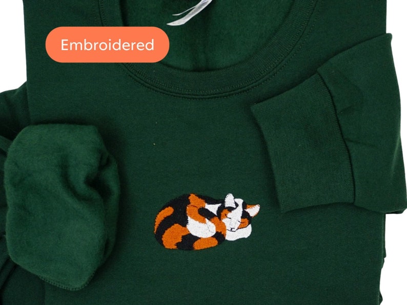May include: A dark green sweatshirt with an embroidered image of a calico cat sleeping. The sweatshirt has the word "Embroidered" written on it.