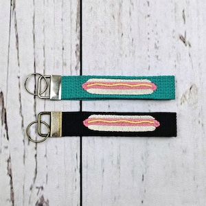 May include: Two fabric keychains featuring embroidered hot dog designs. One keychain has a teal strap, and the other has a black strap. Both include a silver-toned metal hardware attachment with a circular split ring for holding keys securely.