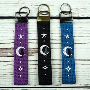 May include: Three fabric keychains in purple, black, and blue. Each keychain features a white embroidered crescent moon with a black cat, stars, and dots. The keychains have metal hardware and are displayed on a light wood surface.