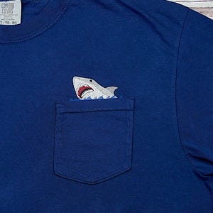 May include: A navy blue t-shirt with a pocket featuring an embroidered shark design. The shark is white and gray with a red mouth, appearing to emerge from the pocket. The shirt is made of a soft material.