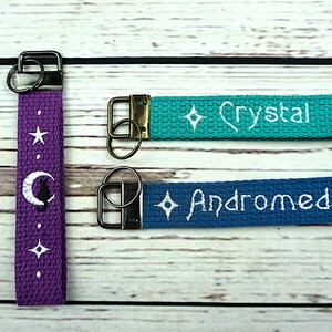 May include: Three keychains with metal hardware. One purple keychain features a white moon and cat design, a star, and dots. The other two are teal and blue, embroidered with the names "Crystal" and "Andromeda" and a diamond.