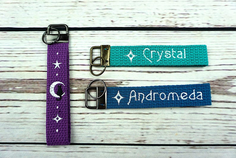 May include: Three keychains with metal hardware. One purple keychain features a white moon and cat design, a star, and dots. The other two are teal and blue, embroidered with the names "Crystal" and "Andromeda" and a diamond.