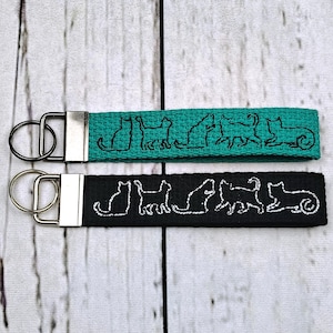 May include: Two fabric keychains with silver-toned metal hardware and split rings. One keychain is teal with black embroidered cat silhouettes, and the other is black with white embroidered cat silhouettes in various poses.
