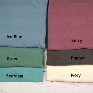 May include: A collection of folded t-shirts in various colors, including ice blue, green, seafoam, berry, pepper, and ivory. Each shirt has a label indicating its color name. The shirts are neatly stacked on a light-colored wooden surface.