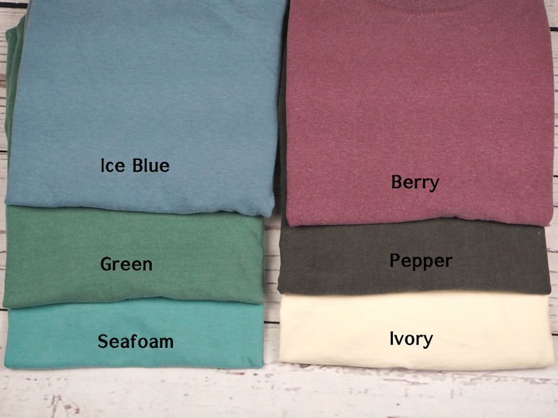 May include: A collection of folded t-shirts in various colors, including ice blue, green, seafoam, berry, pepper, and ivory. Each shirt has a label indicating its color name. The shirts are neatly stacked on a light-colored wooden surface.