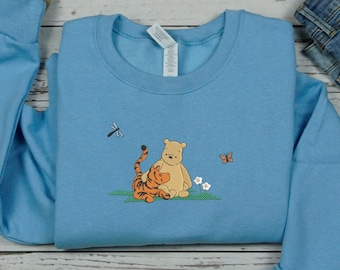 May include: A light blue sweatshirt with an embroidered image of Winnie the Pooh and Tigger hugging. The image is surrounded by a few small flowers and a dragonfly.