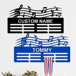 Könnte beinhalten: Ein blauer und schwarzer Medaillenhalter mit einem Musiknoten-Design. Der Halter hat Platz für einen Namen und mehrere Reihen für Medaillen. Der Halter ist an einer weißen Wand montiert, mit zwei Topfpflanzen und einem Glas auf dem Regal darunter.