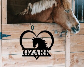 May include: Black metal horse head silhouette inside a heart shape with the word "OZARK" underneath. The heart is hanging on a wooden stall door with a brown horse resting its head on the top of the door.