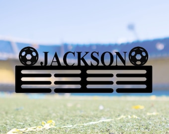 May include: Black metal soccer medal display with the name "JACKSON" and two soccer balls on either side. The display has eight slots for medals.