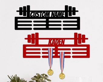 May include: Black and red metal medal display rack with a barbell design. The rack has slots for medals and a space for a name. The name "Kaden" is displayed on the red rack.