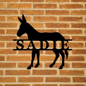 May include: Black metal silhouette of a donkey with the name "SADIE" cut out in the middle of the animal. The silhouette is mounted on a brick wall.