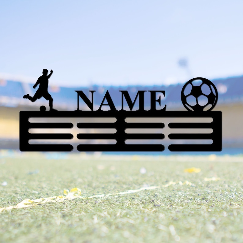 May include: A black metal medal hanger featuring a silhouette of a soccer player kicking a ball on the left, the word NAME in the center, and a soccer ball on the right. The hanger includes two horizontal rows of slots for displaying medals.