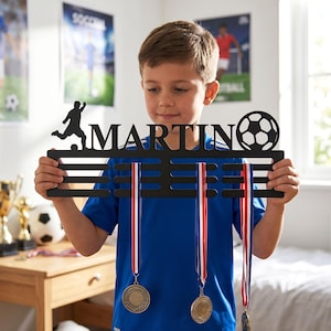 May include: A black metal medal display hanger featuring the name MARTIN in capital letters, a silhouette of a soccer player, and a soccer ball graphic. The hanger includes horizontal bars for hanging medals with ribbons attached.