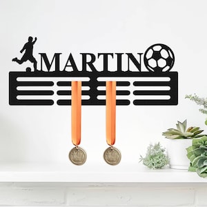 May include: A black metal wall-mounted medal hanger featuring the name MARTIN in capital letters, a silhouette of a soccer player, and a soccer ball graphic. The display includes horizontal slots for hanging two orange-ribboned medals.
