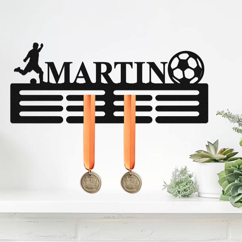 May include: A black metal wall-mounted medal hanger featuring the name MARTIN in capital letters, a silhouette of a soccer player, and a soccer ball graphic. The display includes horizontal slots for hanging two orange-ribboned medals.