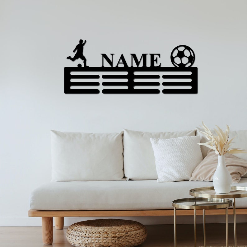 May include: A black wall-mounted metal display rack featuring a silhouette of a soccer player kicking a ball, the word NAME in capital letters, and a soccer ball graphic. The rack includes multiple horizontal slots for hanging items.