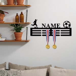 May include: A black metal wall-mounted medal display hanger featuring a silhouette of a soccer player, the word NAME, and a soccer ball graphic. Two gold medals with red, white, and blue ribbons hang from the horizontal display bars.
