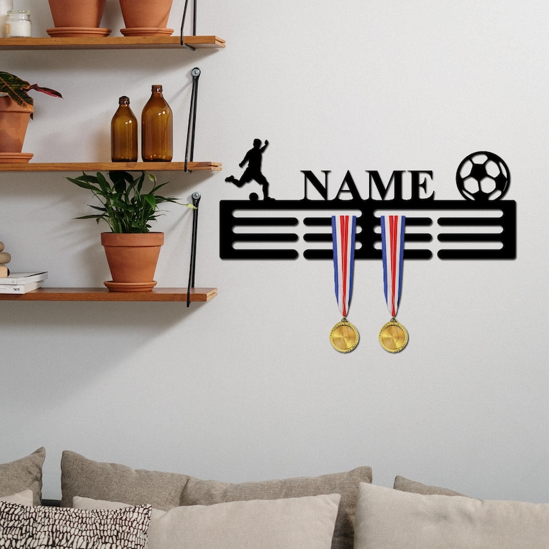 May include: A black metal wall-mounted medal display hanger featuring a silhouette of a soccer player, the word NAME, and a soccer ball graphic. Two gold medals with red, white, and blue ribbons hang from the horizontal display bars.