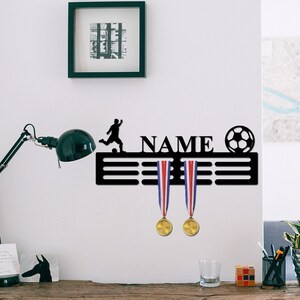 May include: A black metal wall-mounted medal display hanger featuring a silhouette of a soccer player, a soccer ball, and the word NAME in capital letters. Two gold medals with red, white, and blue ribbons hang from the horizontal bars.
