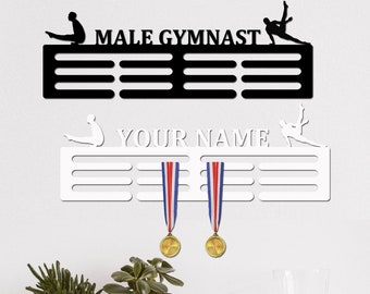 May include: Black and white medal display rack with the text "MALE GYMNAST" and a silhouette of a gymnast. A second white medal rack with the text "YOUR NAME" and a silhouette of a gymnast. Two gold medals are hanging on the white rack.