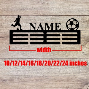 May include: A black metal wall-mounted medal hanger featuring a silhouette of a soccer player kicking a ball, a soccer ball graphic, and the text NAME. The display includes horizontal bars for hanging medals and indicates a width of 10/12/14/16/18/20/22/24 inches.