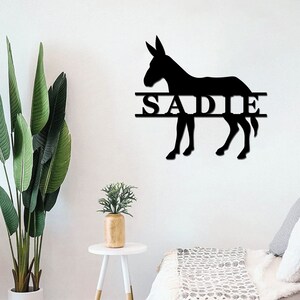 May include: Black metal silhouette of a donkey with the name "SADIE" written across the middle. The silhouette is mounted on a white wall.