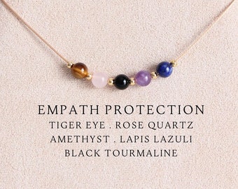 May include: A delicate necklace featuring five small round gemstone beads in tiger eye, rose quartz, black tourmaline, amethyst, and lapis lazuli, separated by tiny gold-tone spacers on a thin cord. Text reads: EMPATH PROTECTION TIGER EYE . ROSE QUARTZ AMETHYST . LAPIS LAZULI BLACK TOURMALINE.