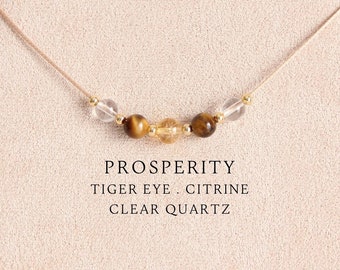 May include: A delicate necklace featuring a thin beige cord strung with two clear quartz beads, two brown tiger eye beads, and one central yellow citrine bead, accented by small gold-tone spacers. Text reads: PROSPERITY TIGER EYE . CITRINE CLEAR QUARTZ.