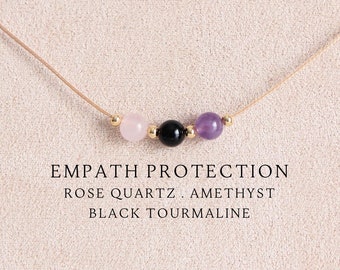 May include: A delicate necklace featuring a thin beige cord with three central beads: a pale pink rose quartz, a glossy black tourmaline, and a translucent purple amethyst, accented by small gold-toned spacers. Text reads: EMPATH PROTECTION ROSE QUARTZ . AMETHYST BLACK TOURMALINE.