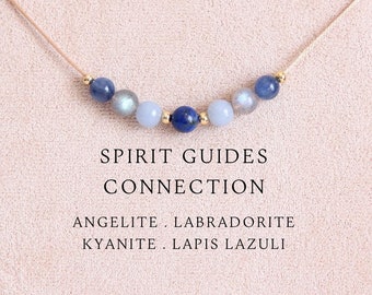 May include: A delicate necklace featuring a thin cord strung with seven small round beads in shades of blue, grey, and gold, including lapis lazuli, labradorite, kyanite, and angelite. The text reads SPIRIT GUIDES CONNECTION ANGELITE . LABRADORITE KYANITE . LAPIS LAZULI.