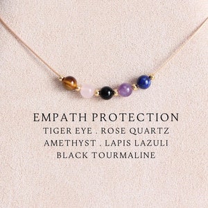 May include: A delicate necklace featuring five small round gemstone beads in tiger eye, rose quartz, black tourmaline, amethyst, and lapis lazuli, separated by tiny gold-tone spacers on a thin cord. Text reads: EMPATH PROTECTION TIGER EYE . ROSE QUARTZ AMETHYST . LAPIS LAZULI BLACK TOURMALINE.
