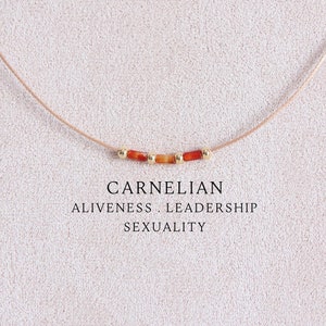 May include: A delicate necklace featuring a thin cord with three small orange carnelian tube beads separated by two tiny gold-toned metallic spheres. The text CARNELIAN ALIVENESS . LEADERSHIP SEXUALITY is printed below the necklace on the display.