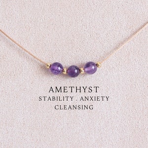 May include: A delicate necklace featuring three round purple amethyst beads separated by small gold-toned metal spacers on a thin beige cord. The text AMETHYST STABILITY . ANXIETY CLEANSING is printed in black below the beads on the display card.