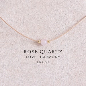 Puede incluir: Un collar delicado con una cuenta de cuarzo rosa pálido centrada entre dos pequeñas esferas metálicas doradas en un cordón beige fino. El texto ROSE QUARTZ LOVE . HARMONY TRUST está impreso en negro debajo de la cuenta en el expositor.