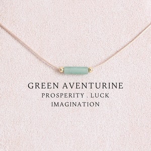 May include: A delicate green aventurine stone necklace on a beige cord with gold accents. The stone is known for its association with prosperity, luck, and imagination.  "GREEN AVENTURINE PROSPERITY. LUCK IMAGINATION" is written below the necklace.