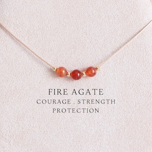 Puede incluir: Un collar delicado con tres cuentas redondas de ágata de fuego de color rojo anaranjado, separadas por pequeños espaciadores dorados en un cordón beige fino. El texto FIRE AGATE COURAGE, STRENGTH PROTECTION está impreso en negro debajo.