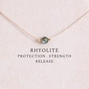 May include: A delicate necklace featuring a single, round rhyolite stone pendant. The stone is a mix of green and gray hues, suspended on a thin, beige cord. The text "RHYOLITE PROTECTION. STRENGTH. RELEASE" is displayed below the necklace.