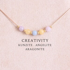 May include: A delicate necklace featuring five small, smooth, round beads in pastel shades of lavender, yellow, and light blue, separated by tiny gold-toned spacers on a thin cord. The text CREATIVITY KUNZITE . ANGELITE ARAGONITE is printed below.