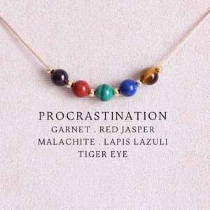 Könnte beinhalten: Eine Halskette mit fünf runden Edelsteinperlen in Dunkellila, Rot, Grün, Blau und Braun, getrennt durch kleine goldfarbene Metallperlen an einer dünnen beigen Kordel. Der Text lautet: PROCRASTINATION, GARNET, RED JASPER, MALACHITE, LAPIS LAZULI, TIGER EYE.