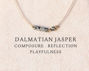 May include: A delicate necklace featuring a thin beige cord, a cylindrical Dalmatian jasper bead, a small round Dalmatian jasper bead, and two tiny gold-toned metallic spacers. The text DALMATIAN JASPER COMPOSURE . REFLECTION PLAYFULNESS is printed below.