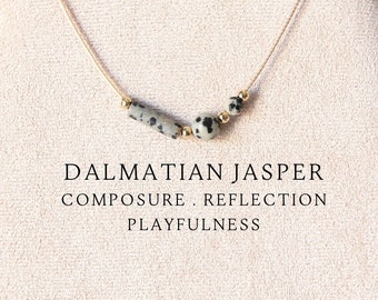 May include: A necklace featuring a thin cord with three Dalmatian jasper beads and small gold-toned spacers. The text DALMATIAN JASPER COMPOSURE . REFLECTION PLAYFULNESS is printed below the necklace on the display stand.