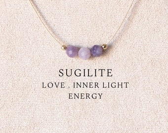 May include: A delicate necklace featuring three small, round, light purple beads centered on a thin metallic chain. Below the necklace, the text reads: SUGILITE, LOVE, INNER LIGHT, ENERGY. The jewelry is displayed on a beige, leaf-shaped stand.
