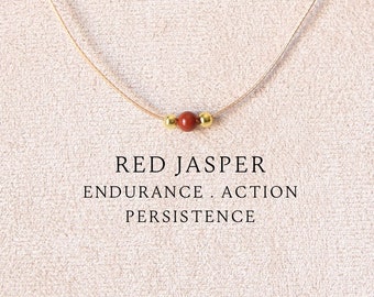 May include: A delicate necklace featuring a single round red jasper bead flanked by two small gold-toned metallic beads on a thin beige cord. The text RED JASPER ENDURANCE . ACTION PERSISTENCE is printed below the bead on the display card.