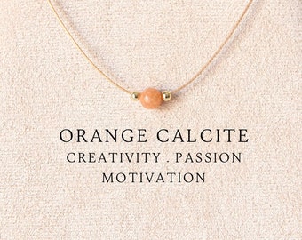 Puede incluir: Un collar delicado con una cuenta redonda de calcita naranja flanqueada por dos pequeños espaciadores metálicos dorados en un cordón beige fino. El texto ORANGE CALCITE CREATIVITY . PASSION MOTIVATION está impreso en negro bajo la cuenta.