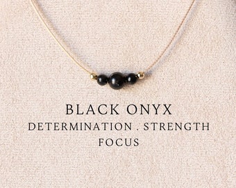 May include: A delicate necklace featuring a thin beige cord and three smooth, round black onyx beads centered with two tiny gold-toned accents. The text BLACK ONYX DETERMINATION . STRENGTH FOCUS is printed below the beads on the display.