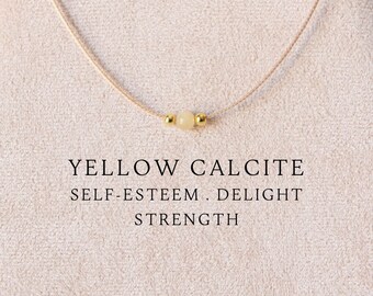 Puede incluir: Un collar delicado con una cuenta única de calcita amarilla pálida flanqueada por dos pequeñas esferas de metal dorado en un cordón beige fino. El texto YELLOW CALCITE SELF-ESTEEM . DELIGHT STRENGTH aparece impreso bajo el collar.